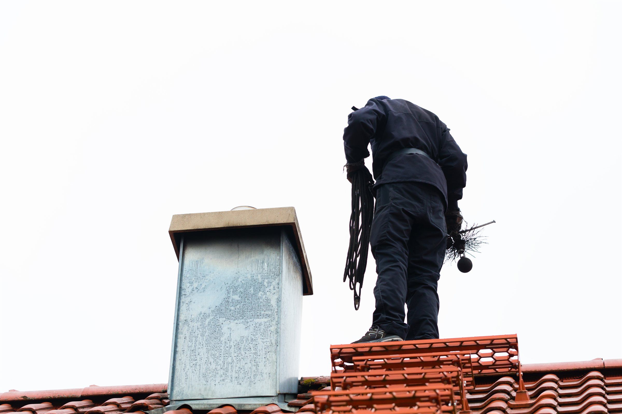 Chimney sweep standing on a tiled roof beside a chimney with cleaning brushes